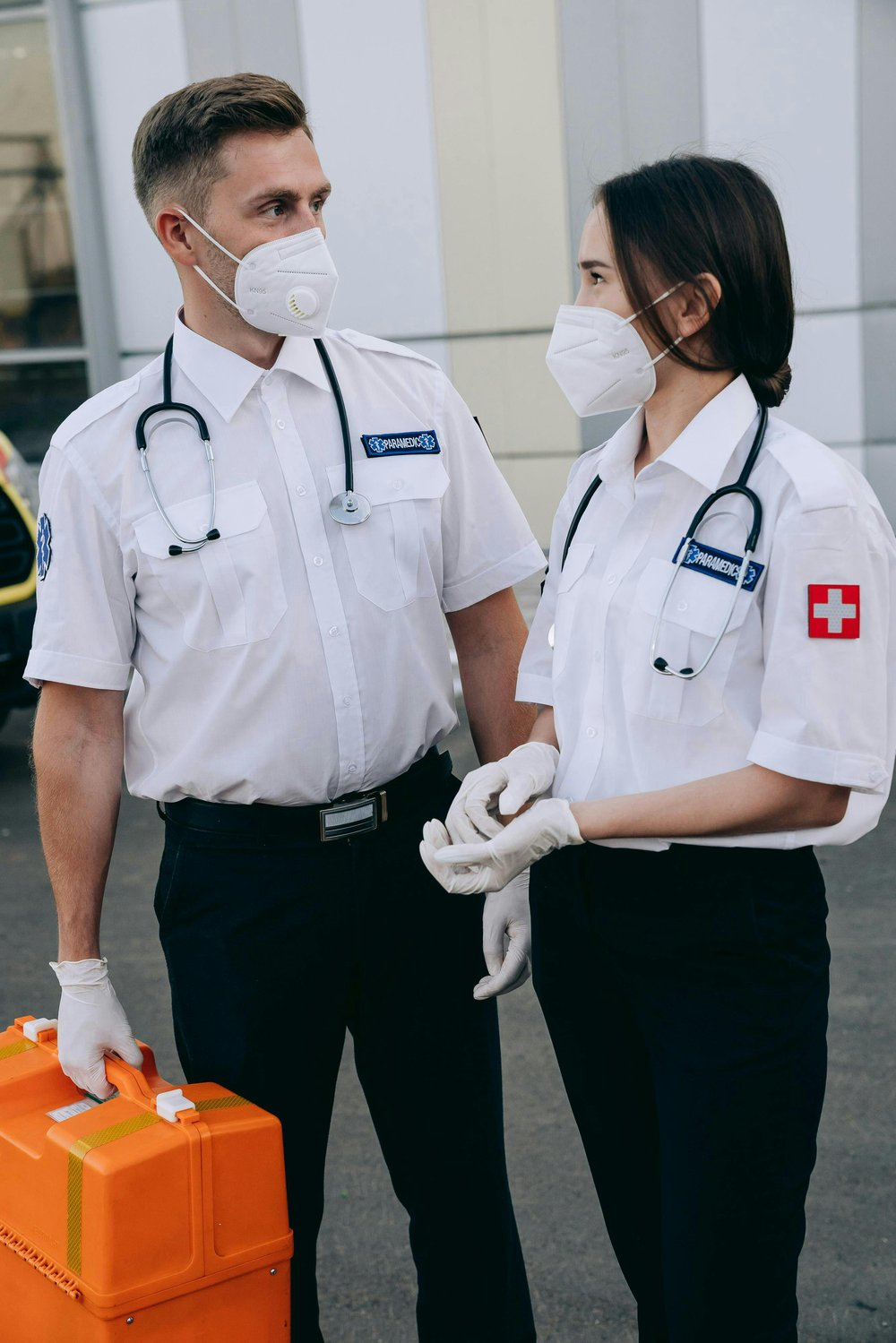 Deux soignants masqués en uniforme blanc, avec stéthoscope, gants et trousse de secours orange.